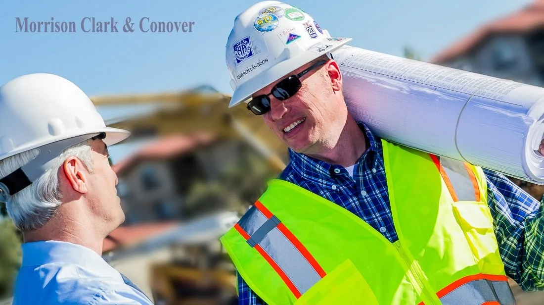 Two construction workers with safety helmets and vests at a construction site, one holding blueprints. Background includes a partly blurred building and clear sky.