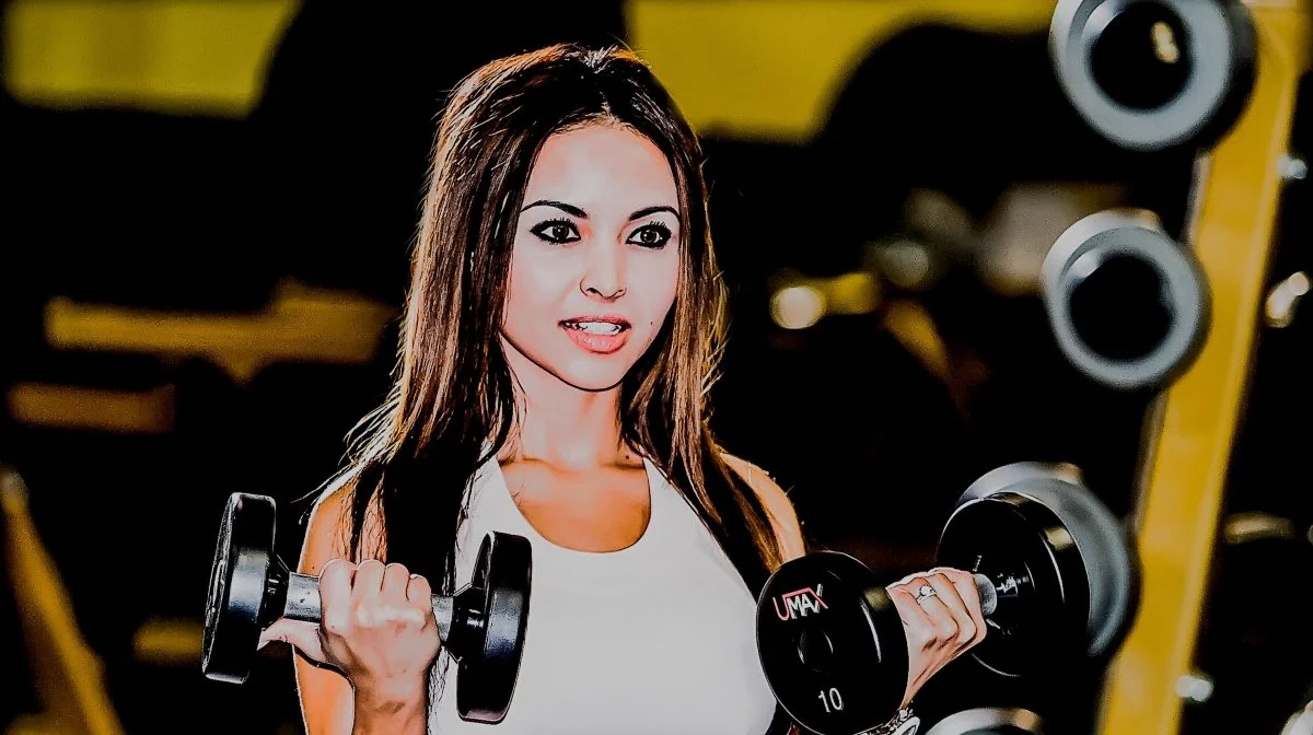 A woman lifting dumbbells in a gym, wearing a white tank top, with a focused expression.