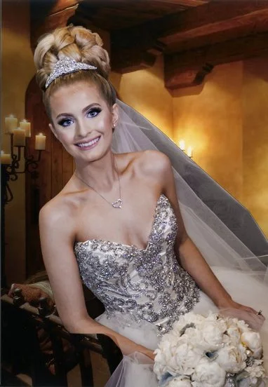 Smiling bride with an elaborate hairstyle, wearing a sparkling wedding gown, and holding a bouquet of white flowers indoors.