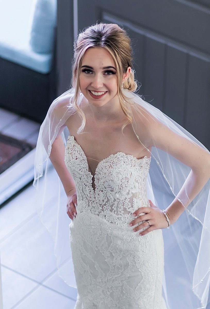 A smiling bride in a white lace wedding gown with a sheer veil, standing indoors near a door with a window.