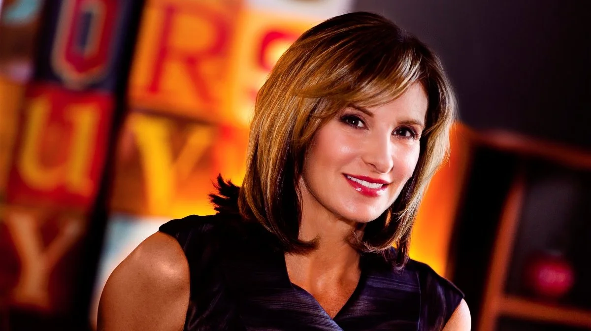 A woman with shoulder-length blond hair smiling, wearing a black sleeveless top, in front of colorful blurred background with yellow and orange hues.