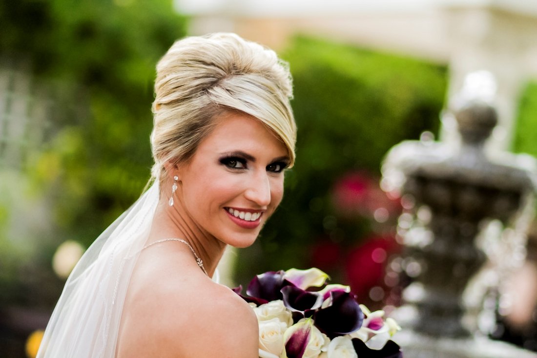 A smiling bride outdoors holding a bouquet of calla lilies and roses, with her hair styled in an elegant updo and wearing jewelry, against a garden background.
