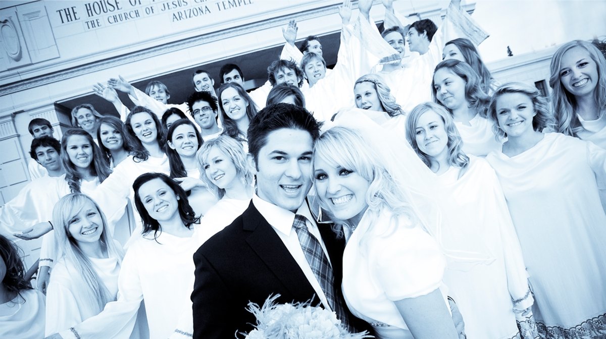 A large group of young people dressed in white robes, celebrating outside a church building, with a man and woman in the foreground smiling at the camera.