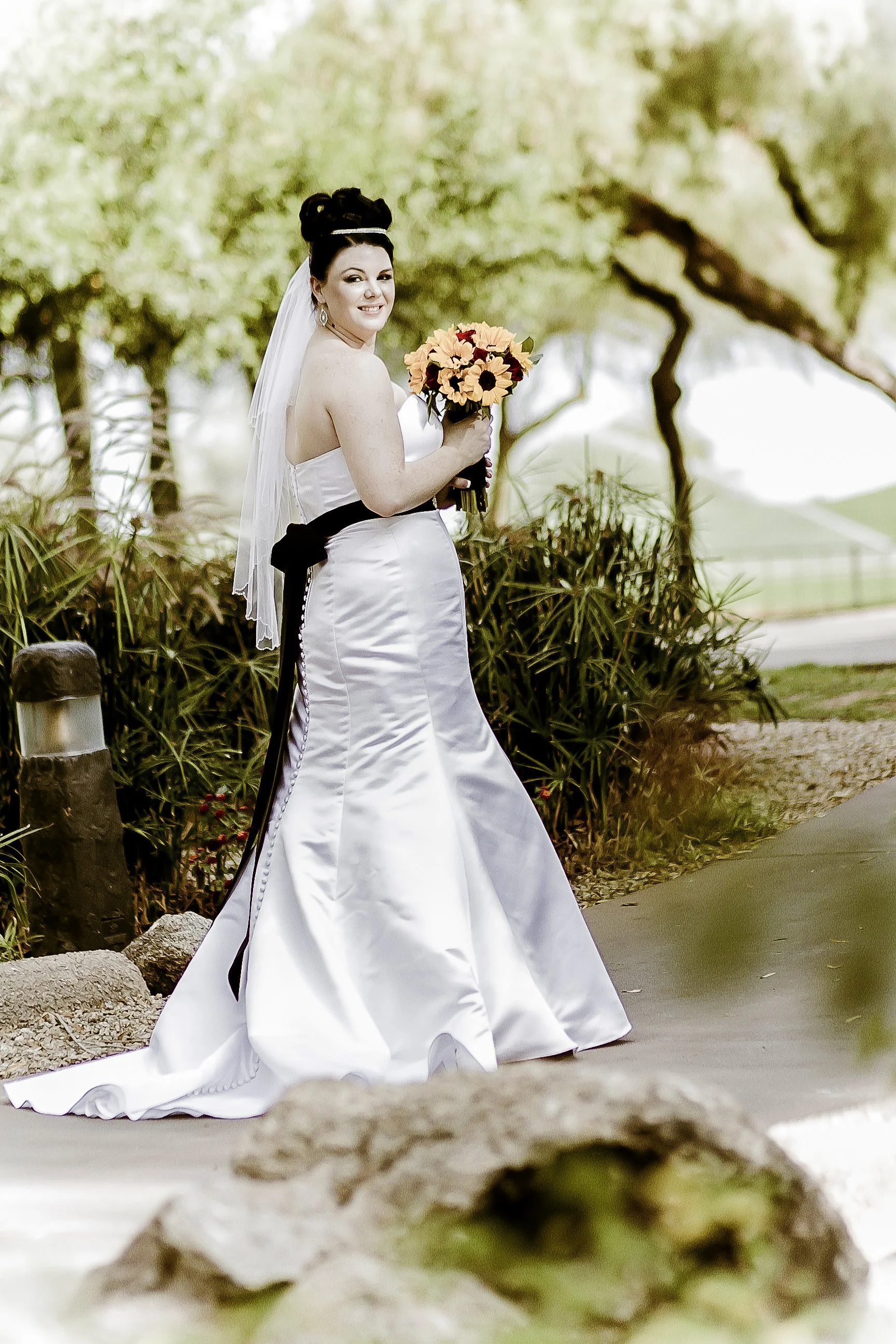 A bride in a white wedding dress standing outdoors on a paved pathway, holding a bouquet of yellow and orange flowers, with greenery and trees in the background.
