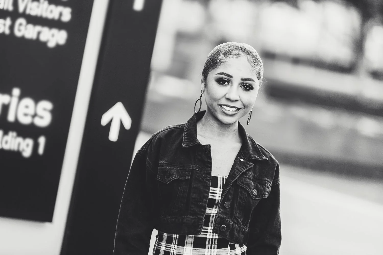 Black and white photo of a young woman smiling outdoors, wearing a denim jacket and hoop earrings, standing near a parking garage sign.