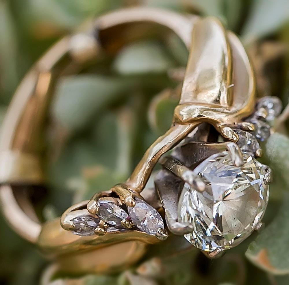 Close-up of a gold ring with a large central diamond and smaller diamonds on the sides, placed on green leaves.