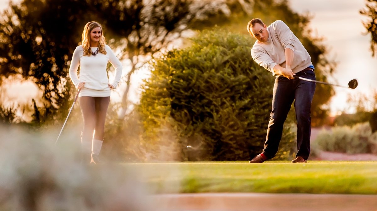 A man and woman playing golf outdoors during sunset, with trees in the background. The man is preparing to hit a golf ball while the woman stands nearby watching.