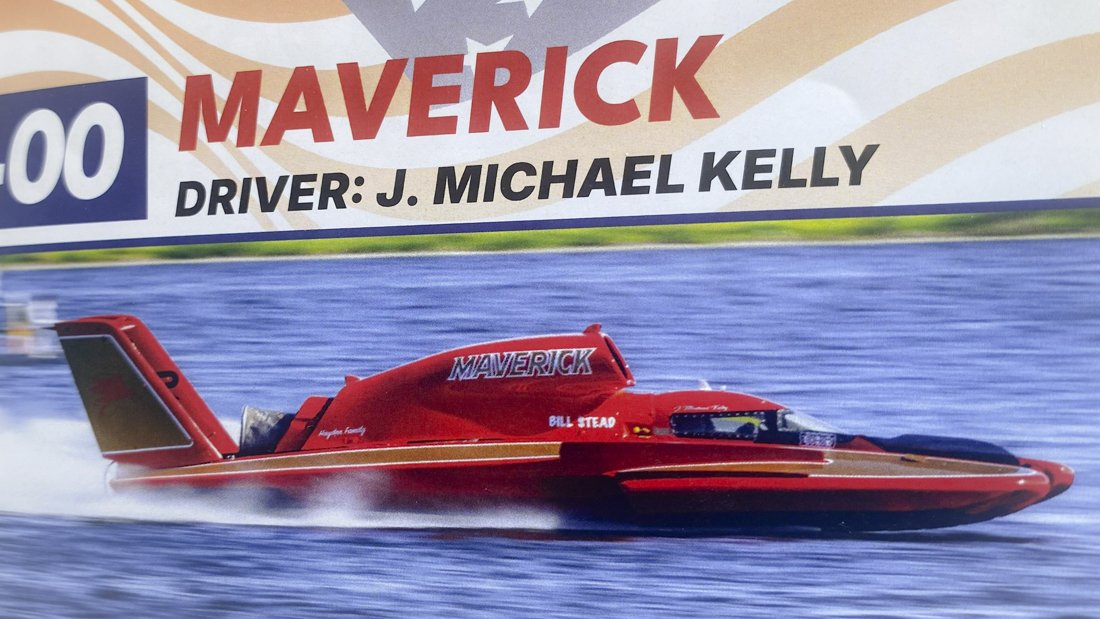 Red race boat with the word "MAVERICK" on top, speeding on water with a spray trail, under a sign that says "MAVERICK" and "DRIVER: J. MICHAEL KELLY".