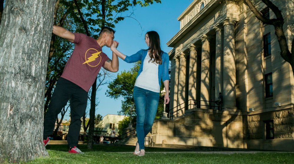 A young couple on a city sidewalk; the man playfully grabs the woman's hand as if shielding her from an imaginary punch, both smiling. The man wears a maroon t-shirt with a yellow lightning bolt symbol and black pants, the woman wears a white top, bl