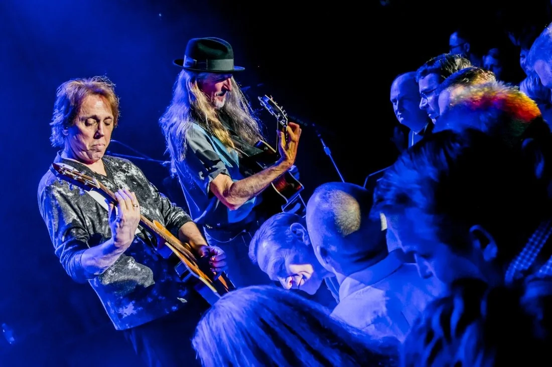 Two musicians playing guitars on stage during a live performance, with seated audience in front of them.