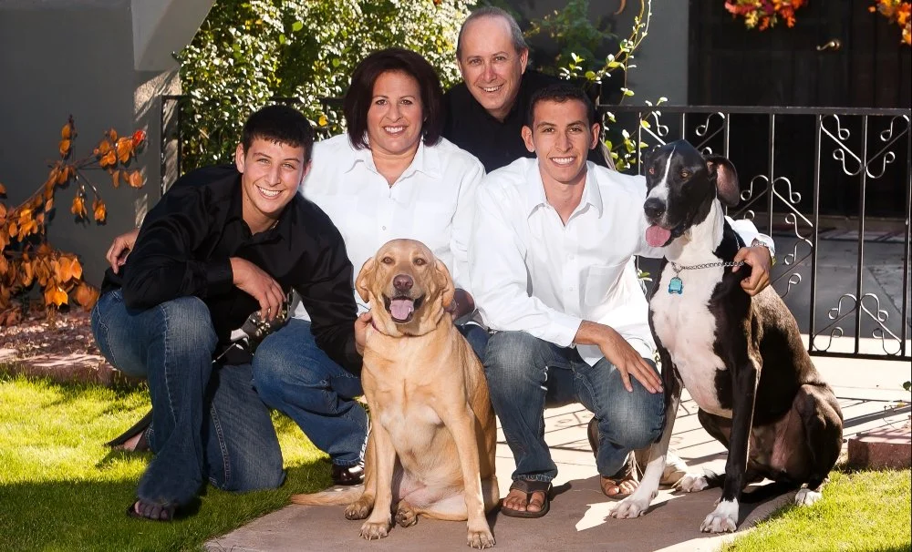 A family of five posing outdoors on a sunny day, with two large dogs and various plants in the background.