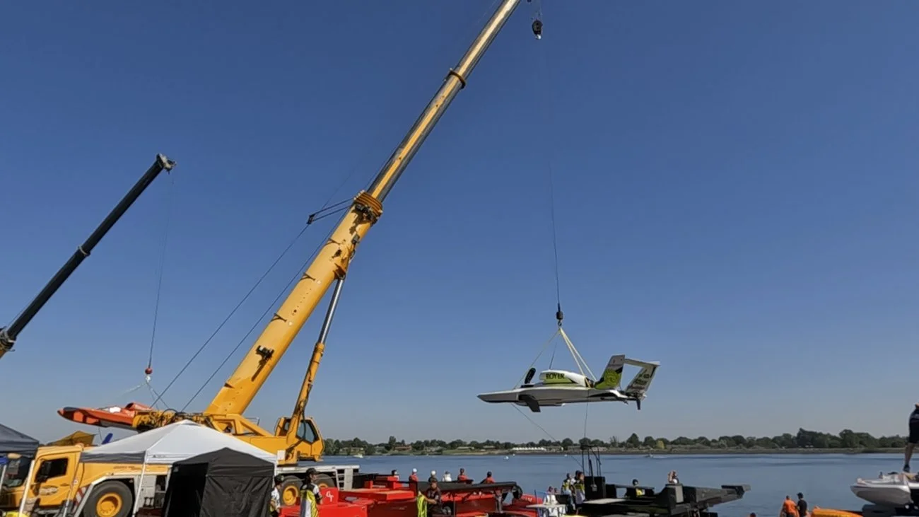 A large crane lifting a small airplane over a body of water during a festival or event with people on the shore.