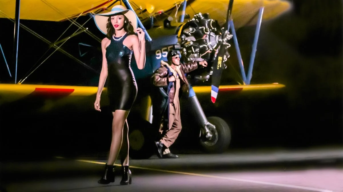 A woman in a black dress and wide-brimmed hat standing next to a vintage airplane at night.