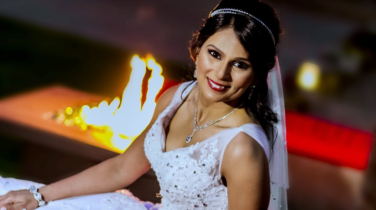 A woman in a white wedding dress and jewelry, smiling at the camera, with a fireplace in the background.