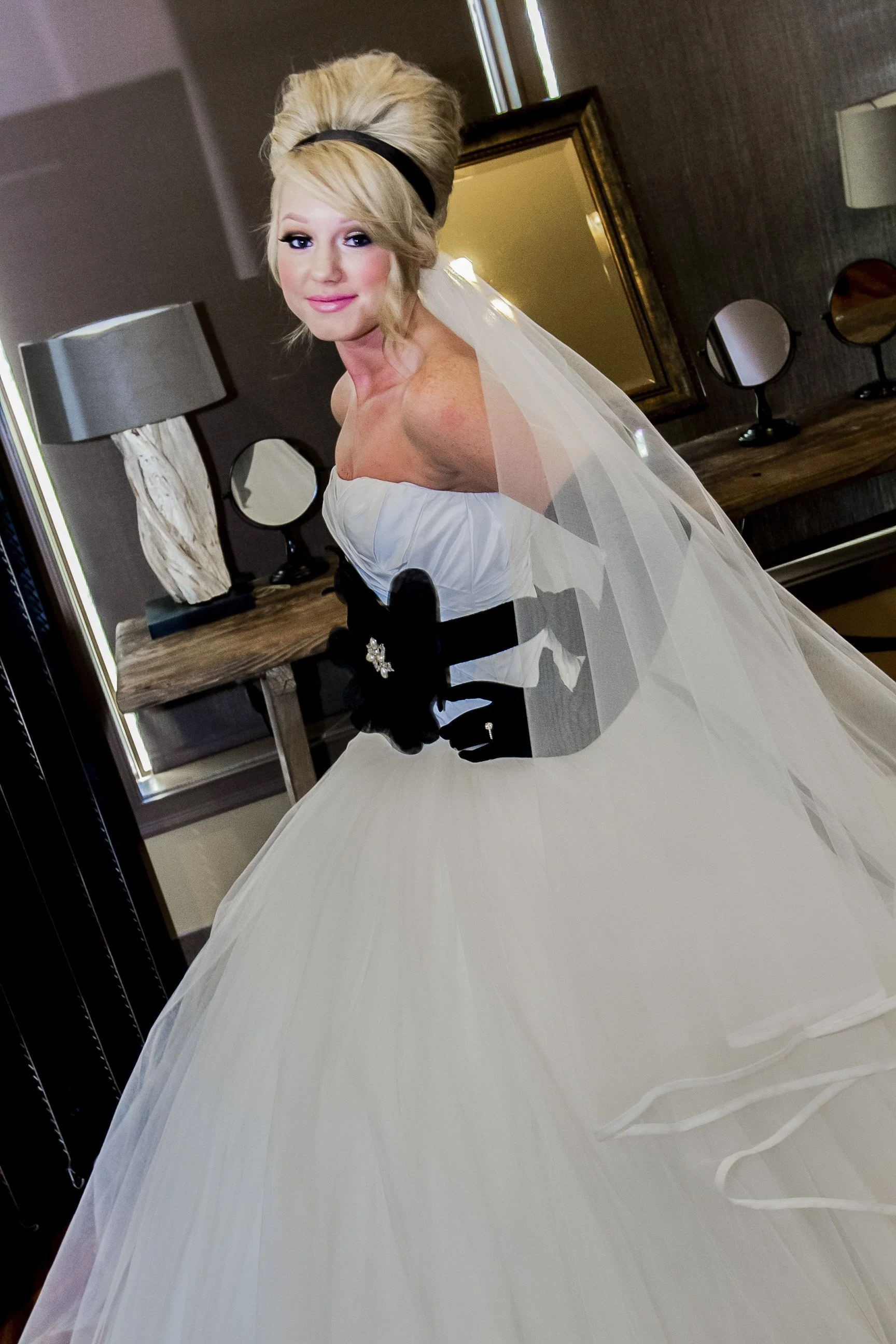 A woman dressed in a wedding gown, with an updo hairstyle, wearing makeup, standing indoors near a mirror, with a table holding two small round mirrors and lamps behind her.