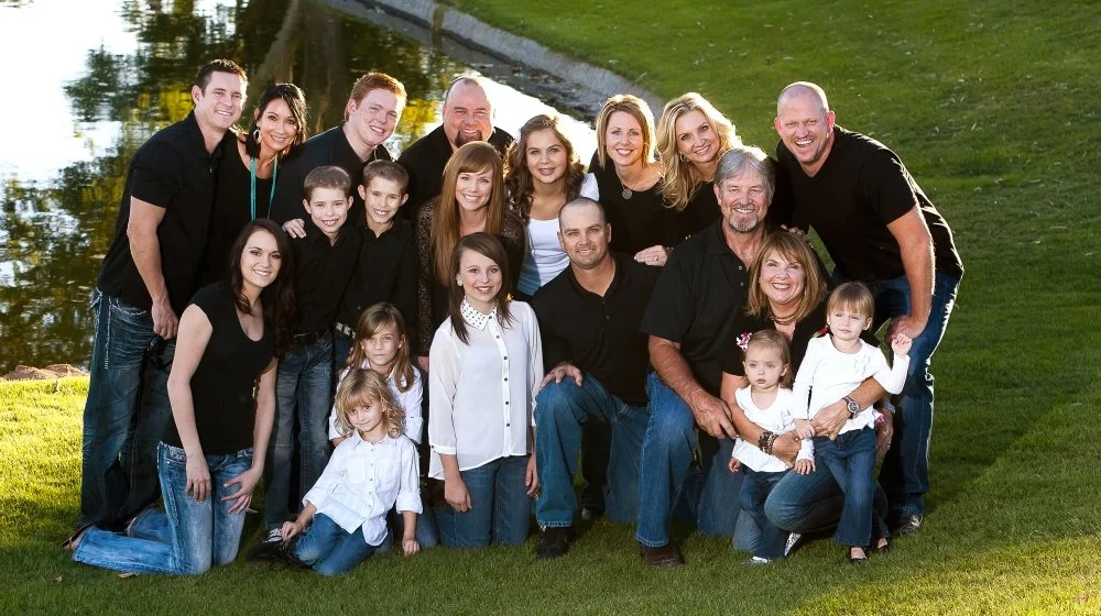 Family group photo outdoors near a pond, with adults, teenagers, and young children smiling and posing together on the grass.