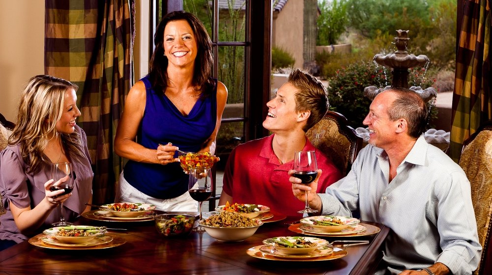 Family gathered around a dining table, smiling, as a woman stands serving food, with outdoor scenery visible through large windows.