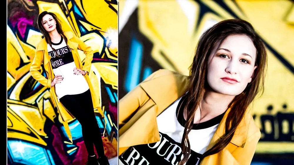 Portrait of a young woman with long brown hair, wearing a yellow jacket and a white and black t-shirt, standing in front of a colorful graffiti wall.