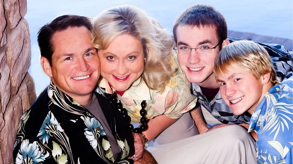 Four smiling people posing closely together outdoors, near a stone wall and body of water in the background.
