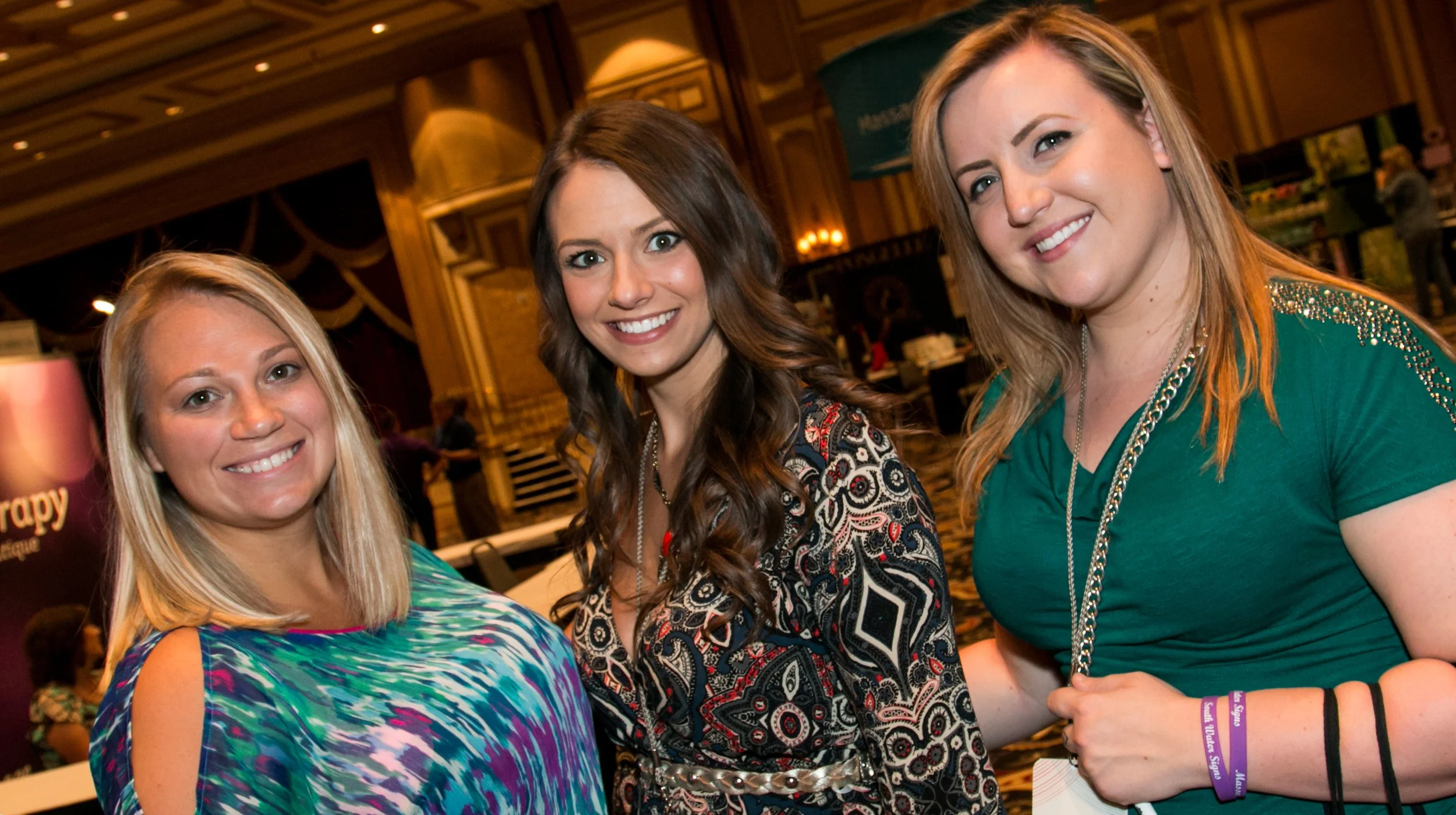 Three women smiling at an indoor event with warm lighting, decorative wood paneling, and informational displays in the background.