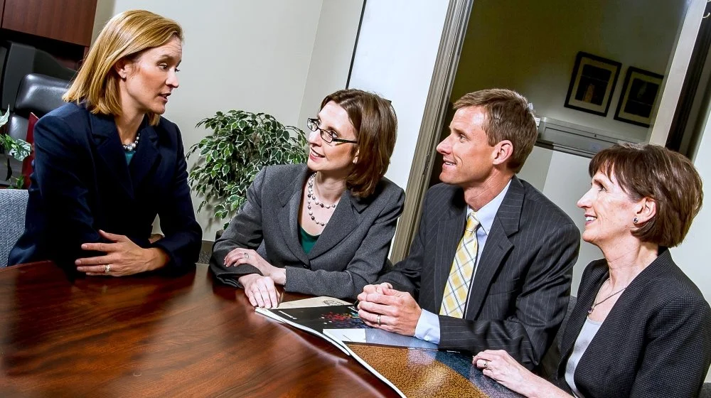 Four business professionals, three women and one man, engaged in a serious discussion in an office conference room, with two women standing and two women and one man sitting around a wooden table.