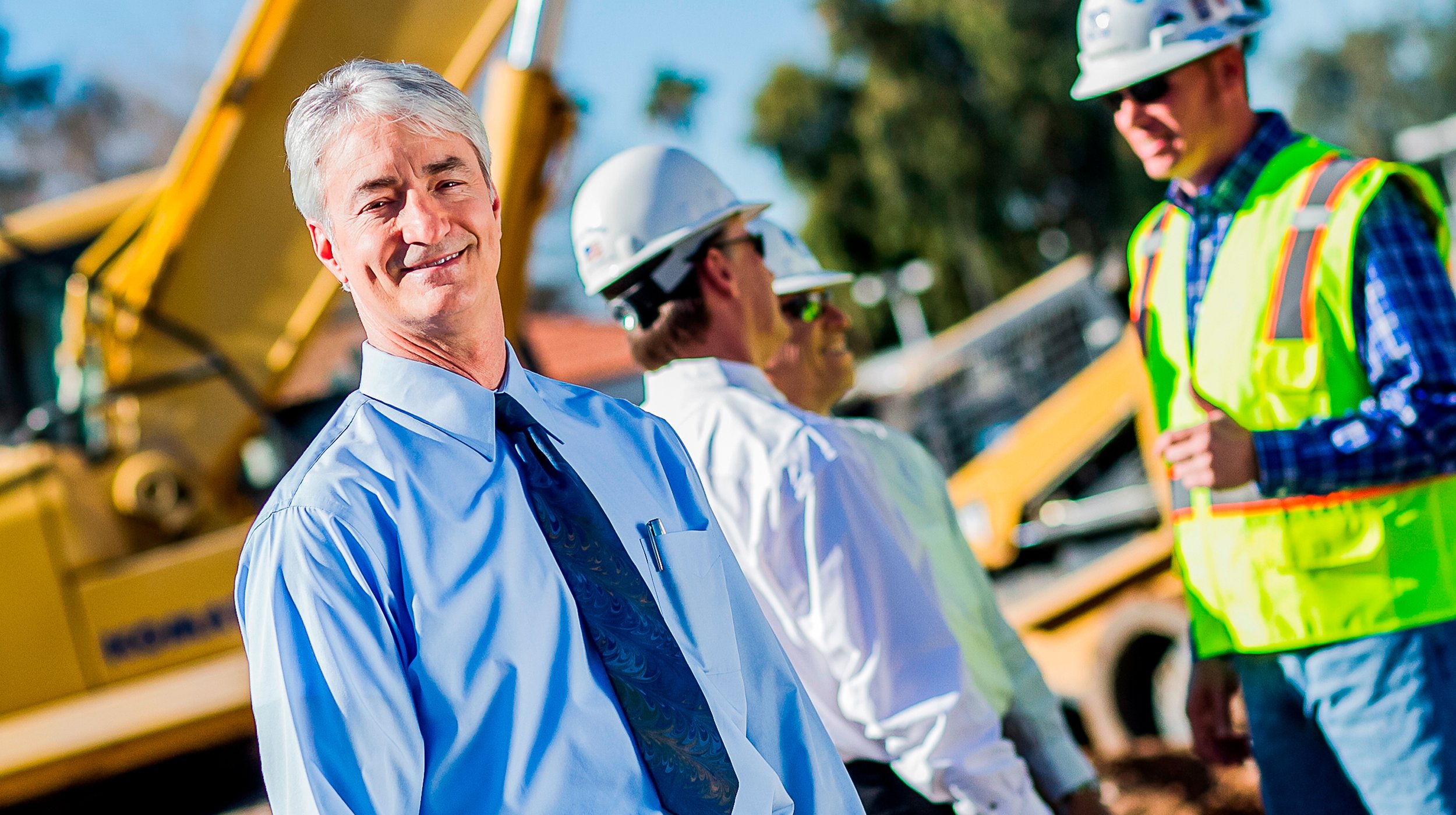 A group of construction professionals outdoors, with a man in business attire smiling in the foreground and workers in safety helmets and uniforms in the background.