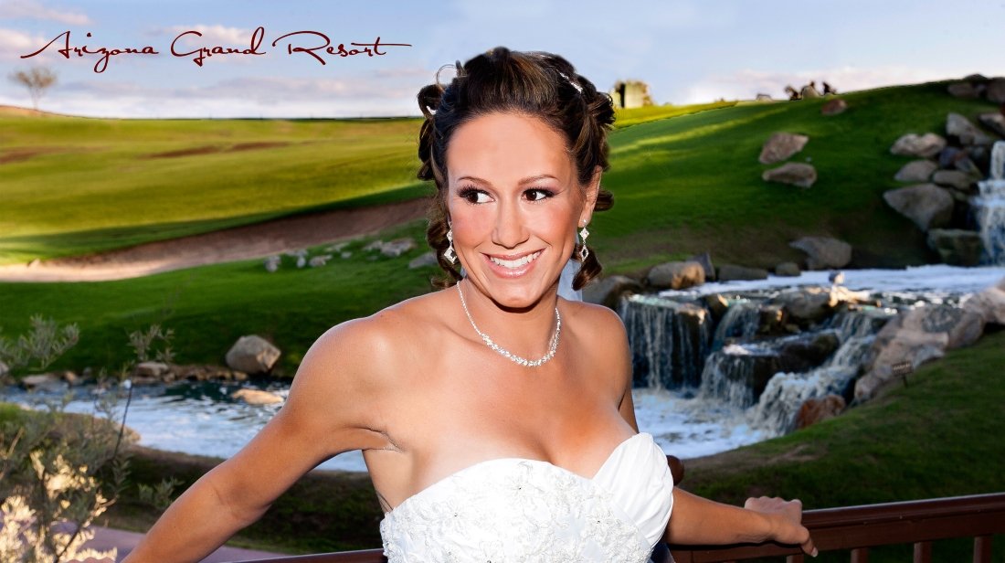 A woman in a white wedding dress smiling on a balcony with a scenic background of green hills, rocks, a waterfall, and a river at Arizona Grand Resort.