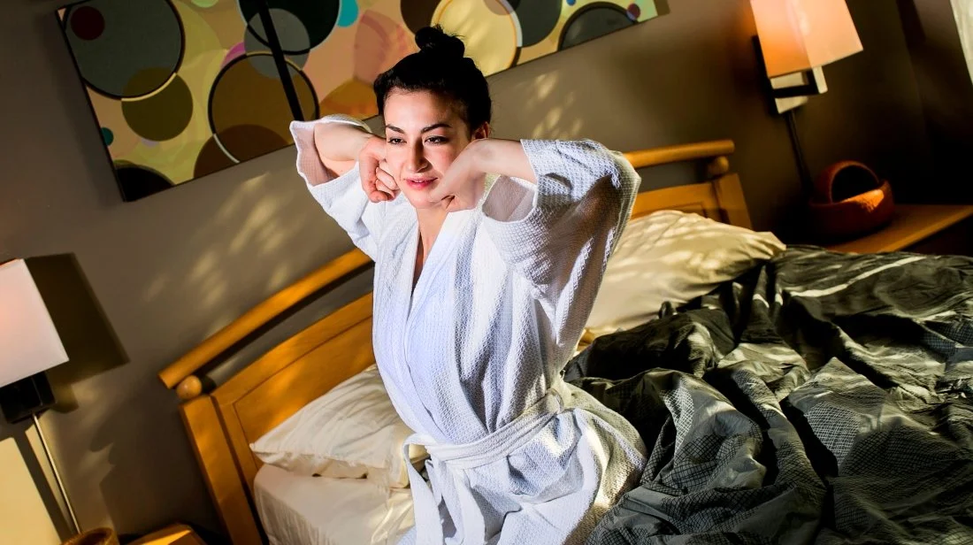 A woman in a white robe stretching in bed in a hotel room.
