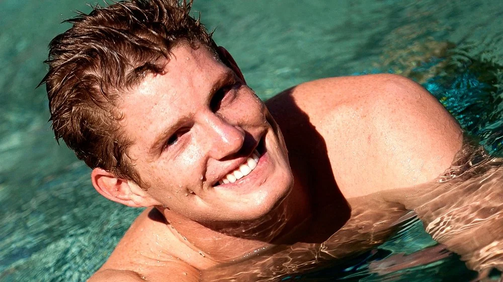 A young man with short, wet hair smiling while swimming in a pool.