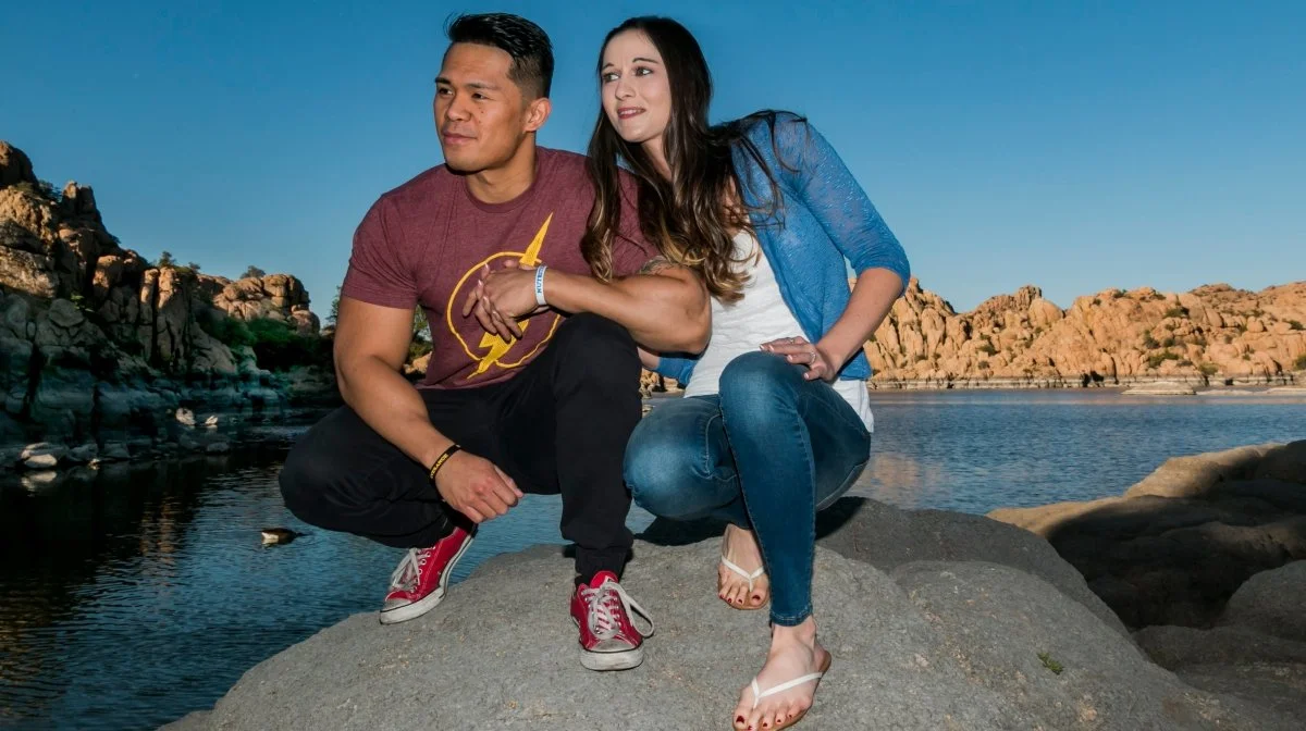 A man and woman squatting on a large rock near a body of water with rocky terrain in the background.