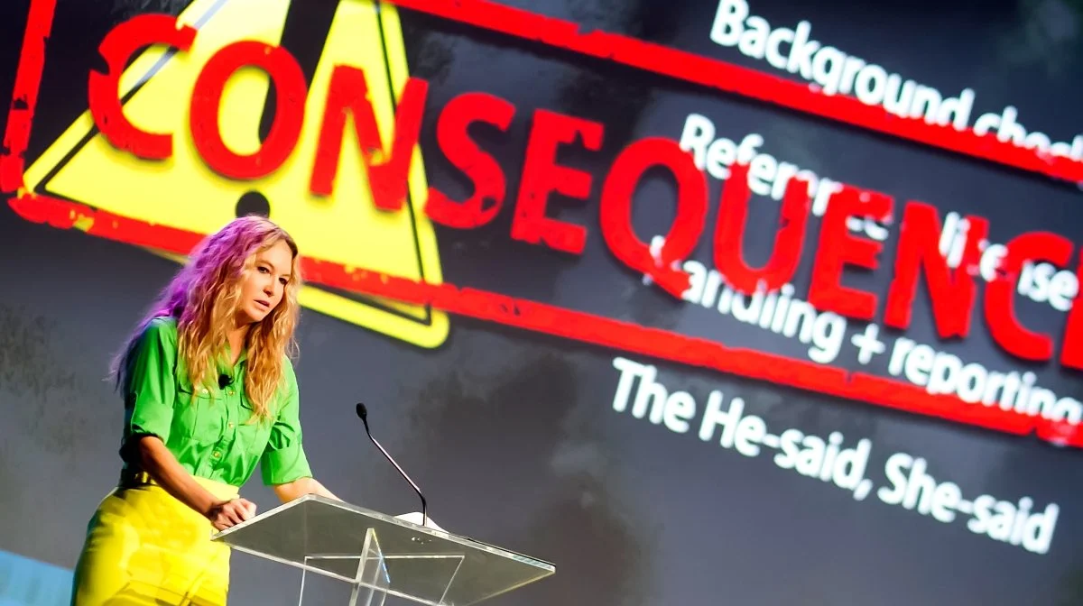 A woman in a bright green shirt and yellow pants speaking at a podium, with a large Halloween-style caution sign background that reads 'CONSEQUENCES' and additional text related to substance use and reporting.