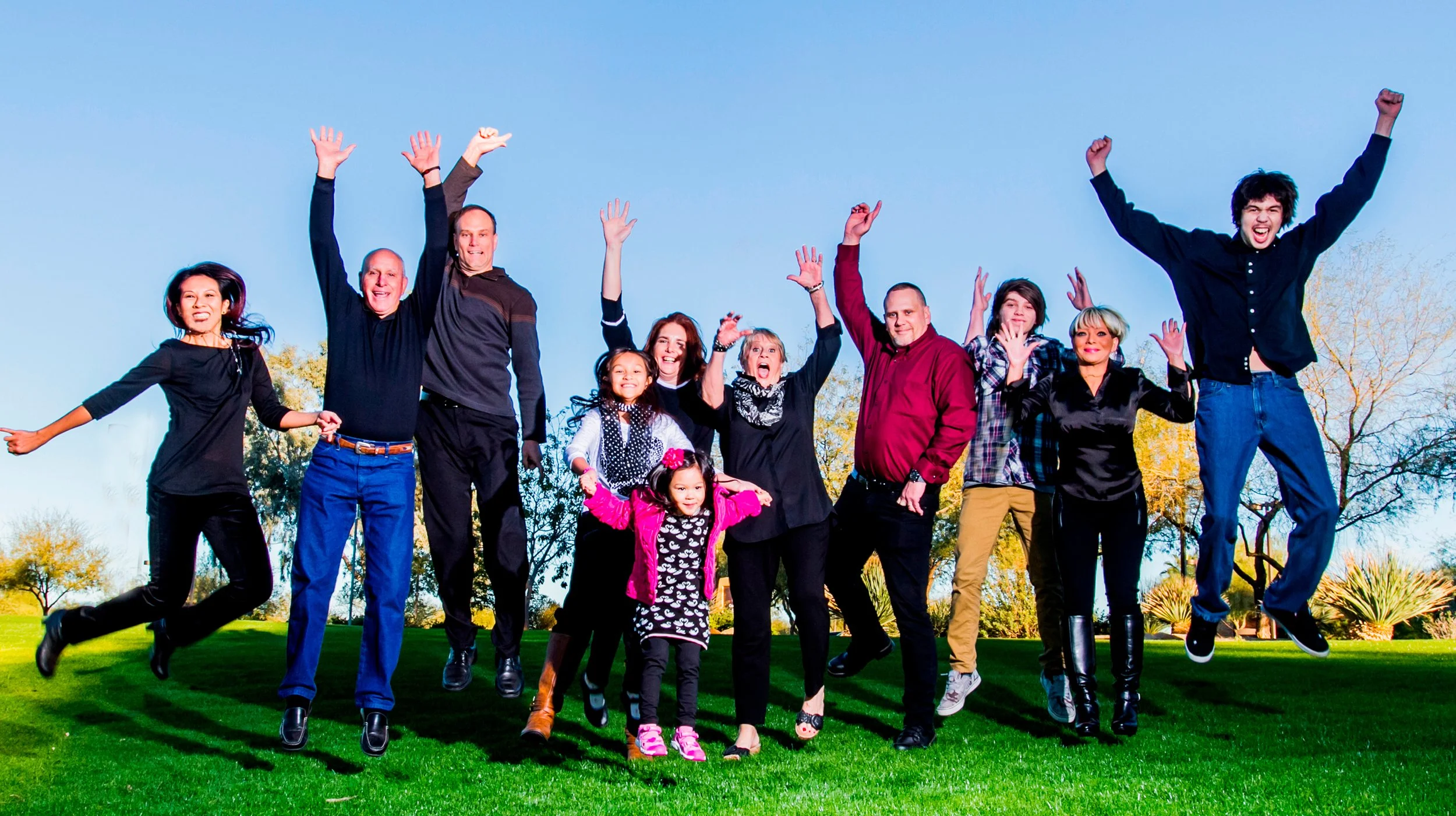 A group of people, including children and adults, jumping in the air outdoors on a grassy field with trees in the background during daytime.