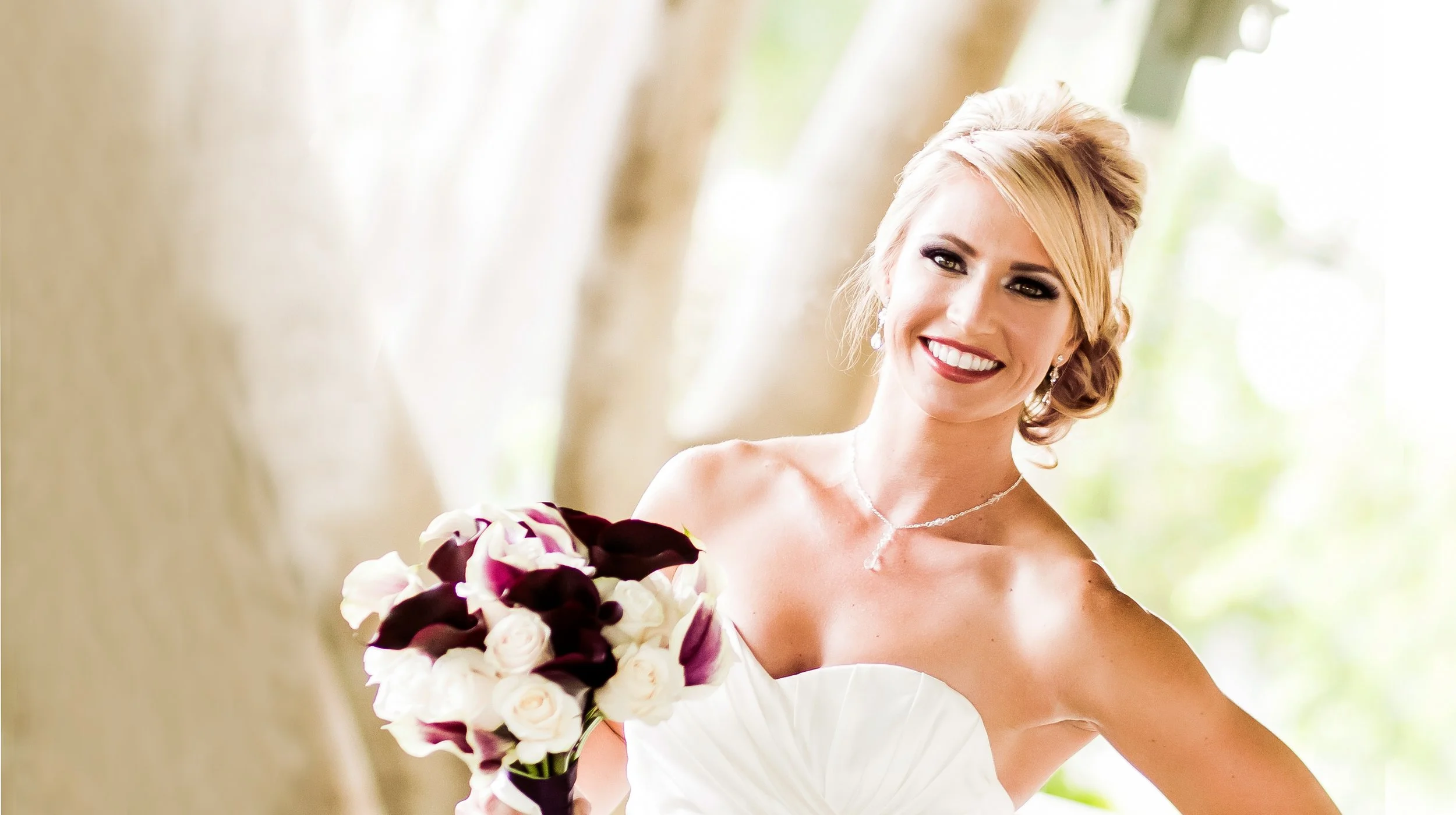 Bride in a white wedding dress holding a bouquet of white and dark purple flowers, smiling in a bright outdoor setting.