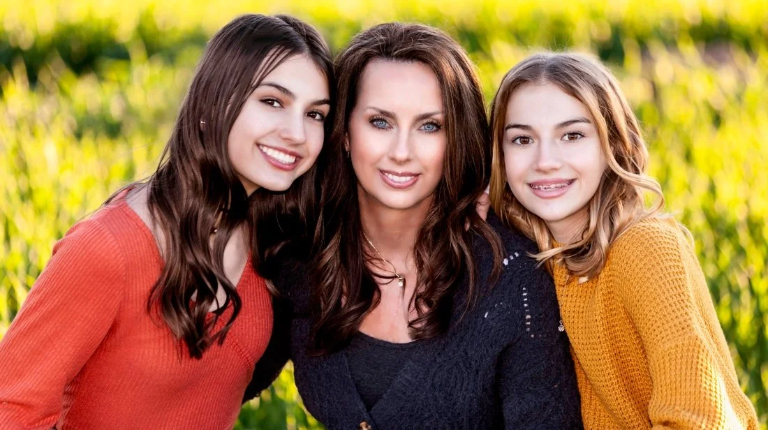 A woman with two young girls outdoors smiling, with a green field in the background.