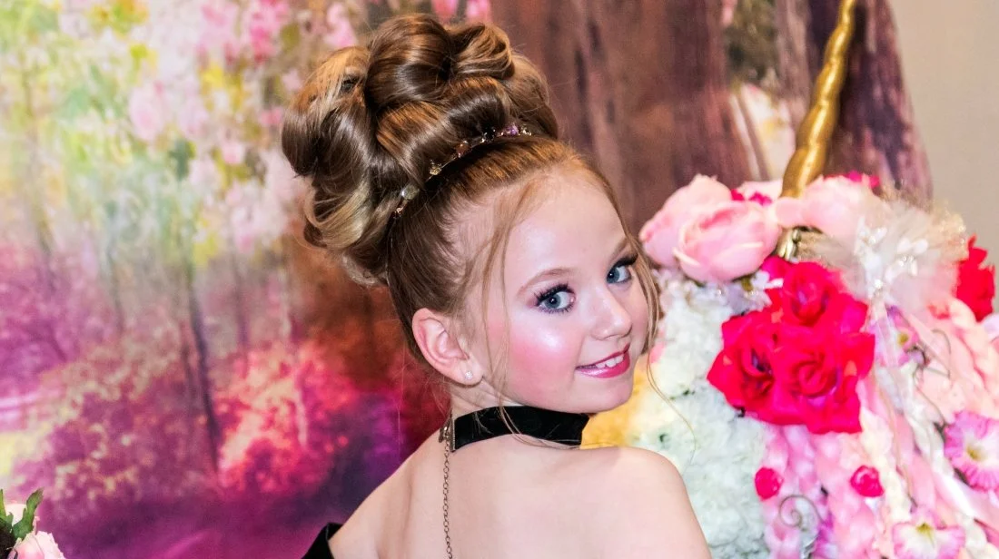 Young girl with styled hair in an updo, wearing a black choker and earrings, smiling at a floral display with pink and white flowers and a golden unicorn horn in the background.