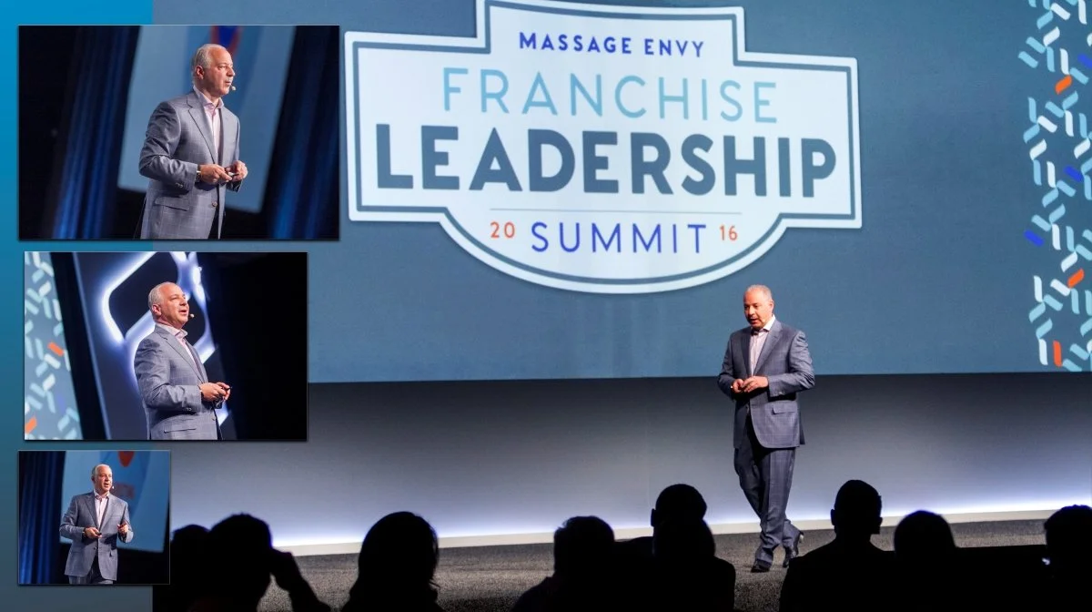 A man in a gray suit presenting at a franchise leadership summit, with a large screen displaying the event logo, which reads 'Massage Envy Franchise Leadership Summit 2016'.