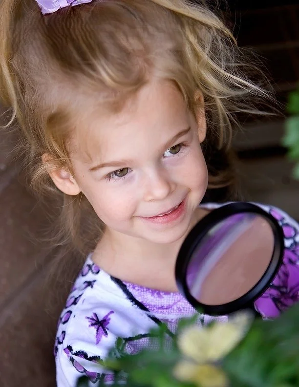 A young girl with light brown hair, wearing a white and purple floral top, holding a magnifying glass and examining plants or flowers outdoors.