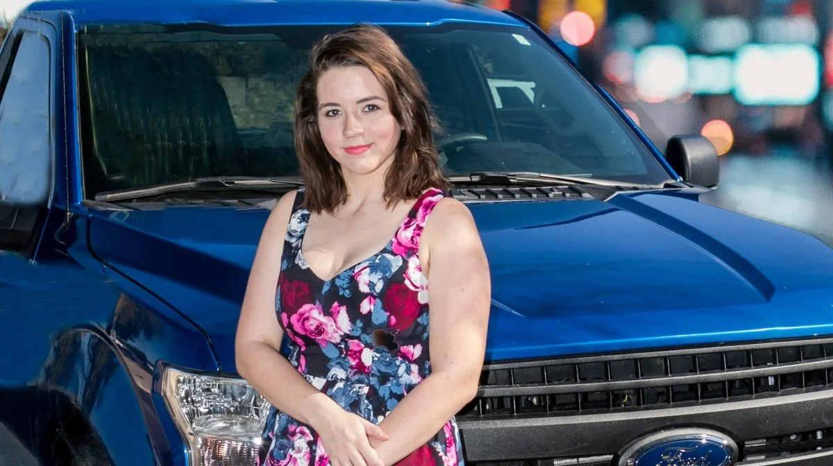 Young woman with shoulder-length brown hair in a floral dress standing in front of a blue pickup truck in an urban street at night.
