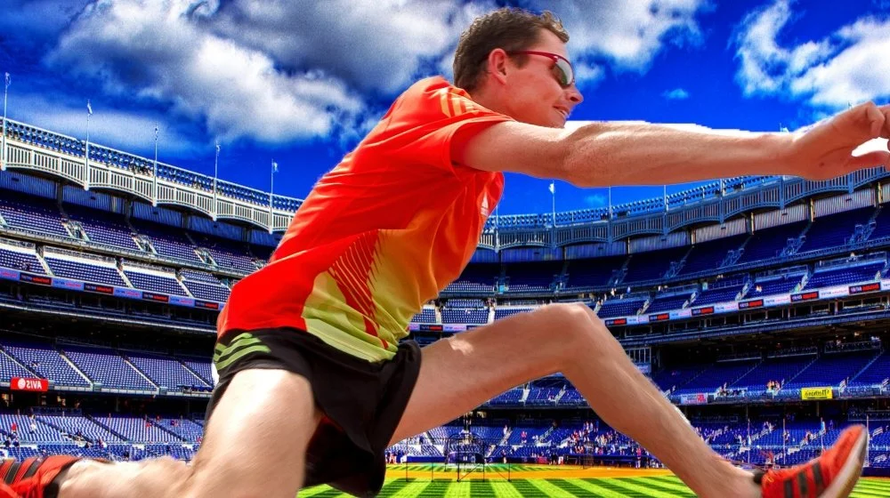 A man in sunglasses and a red and yellow sports shirt is stretching on a tennis court in a large stadium with blue seats, under a partly cloudy sky.