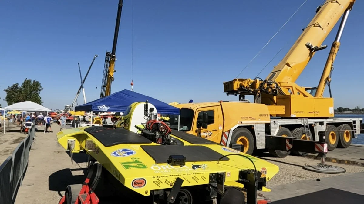 Yellow racing vehicle and a large yellow crane truck on display at an outdoor event, with tents and people in the background under clear blue skies.