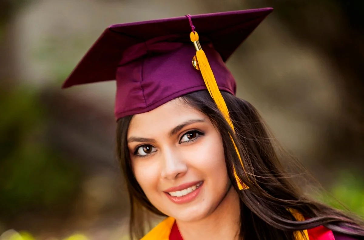 A young woman smiling in her graduation gown and cap with a yellow tassel, outdoors.