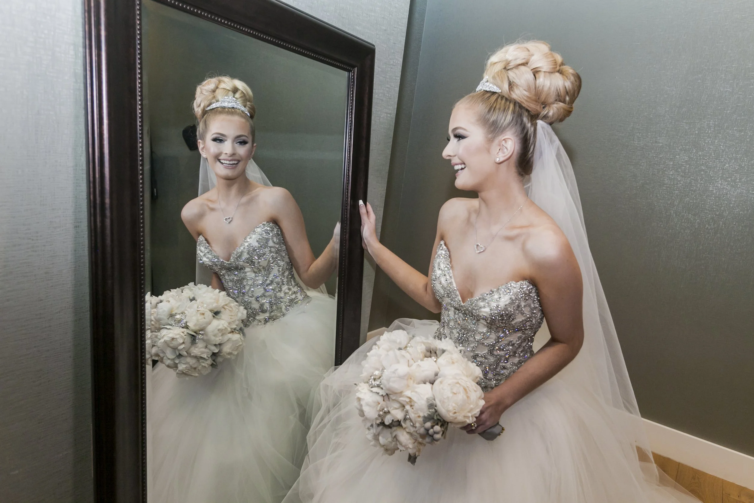 A bride with a flower bouquet looking at her reflection in a mirror, smiling, wearing a detailed sparkling strapless wedding gown and veil, with her hair styled in an elaborate braided updo.