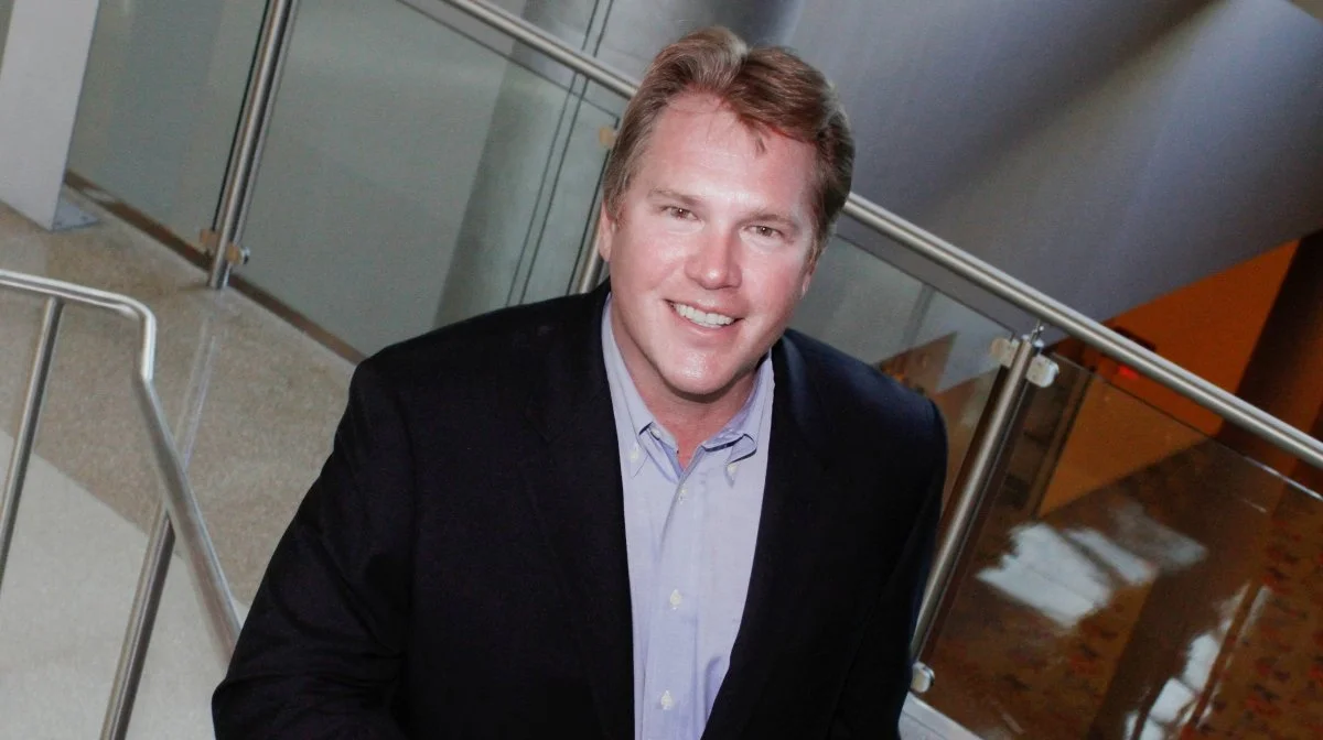 A smiling man with light brown hair wearing a blue dress shirt and black blazer, standing indoors near glass railings.