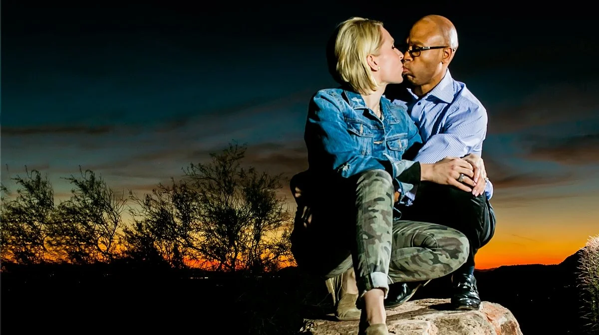 A couple sharing a kiss at sunset, sitting on a rock in a natural outdoor setting with trees and a colorful sky.