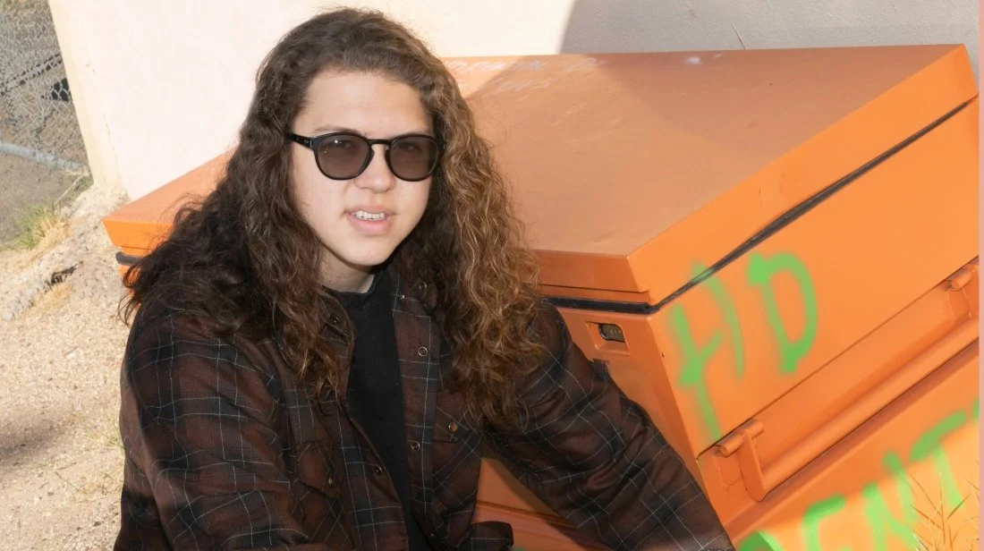 A woman with long curly hair wearing sunglasses and a dark plaid shirt sitting next to an orange dumpster with green graffiti.