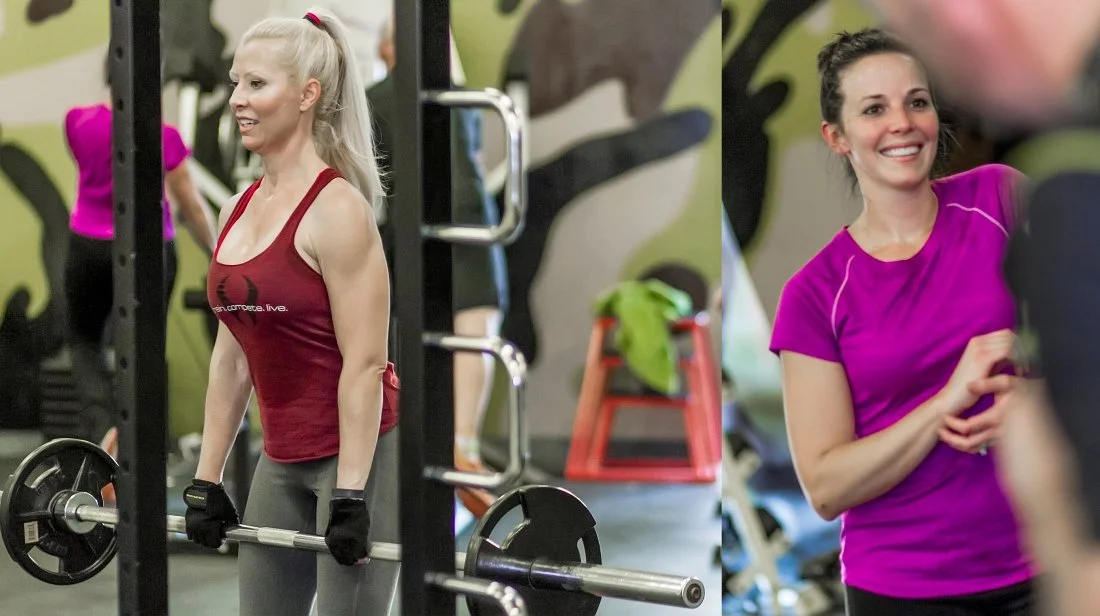 Women working out at a gym, one lifting a barbell, the other smiling and talking, with gym equipment and a green wall in the background.