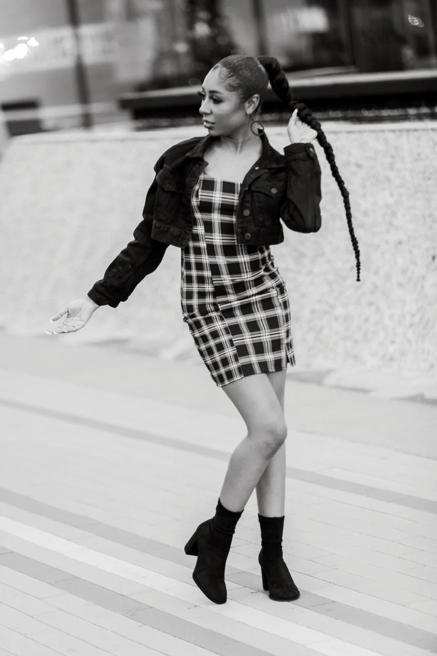 A woman with a long braided ponytail walking on a sidewalk, wearing a plaid dress, black jacket, and high-heeled ankle boots, holding her ponytail with one hand.