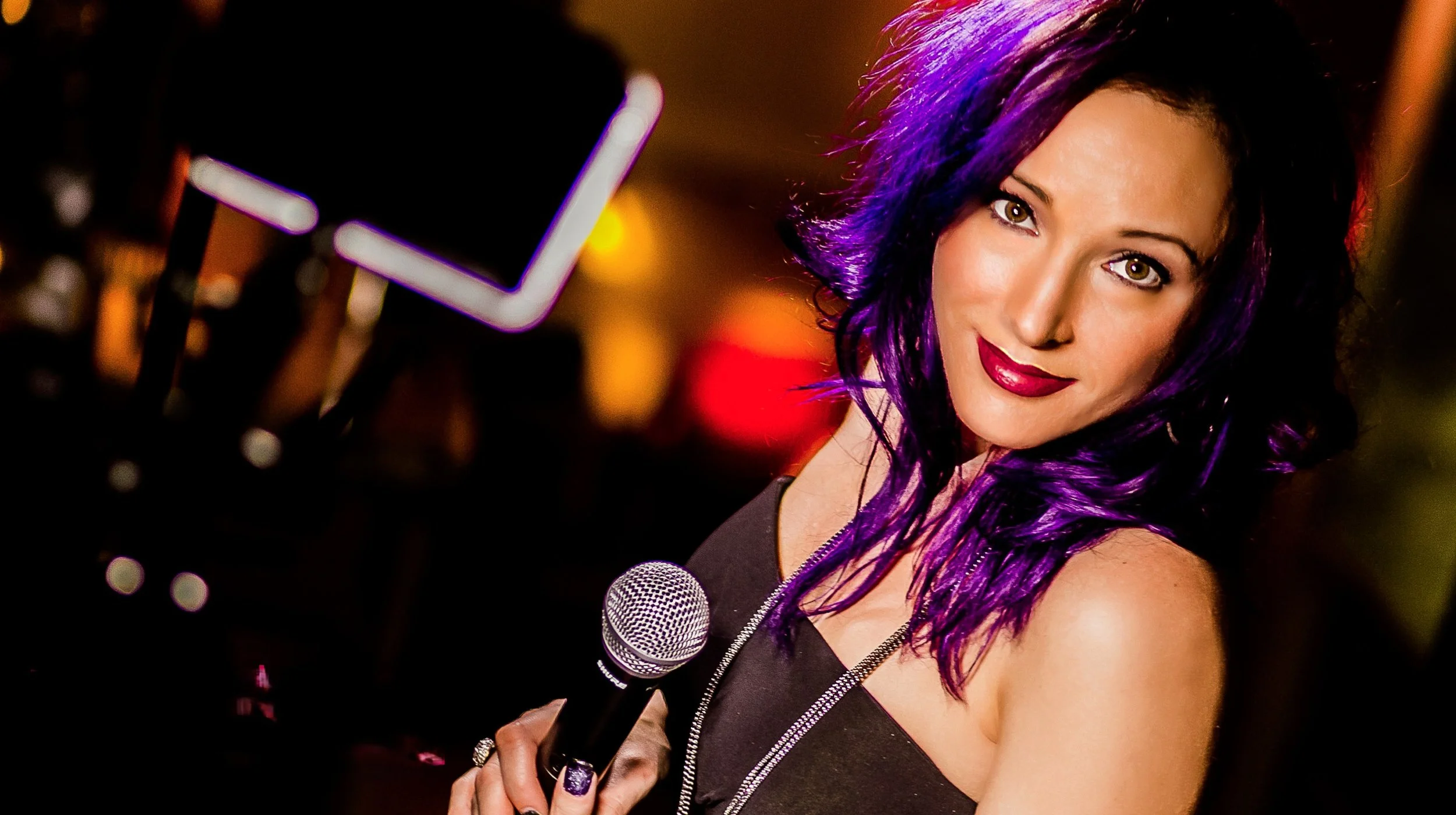 A woman with vibrant purple hair, fair skin, and red lipstick, smiling at the camera while holding a microphone, at an event or party with blurred background lighting.