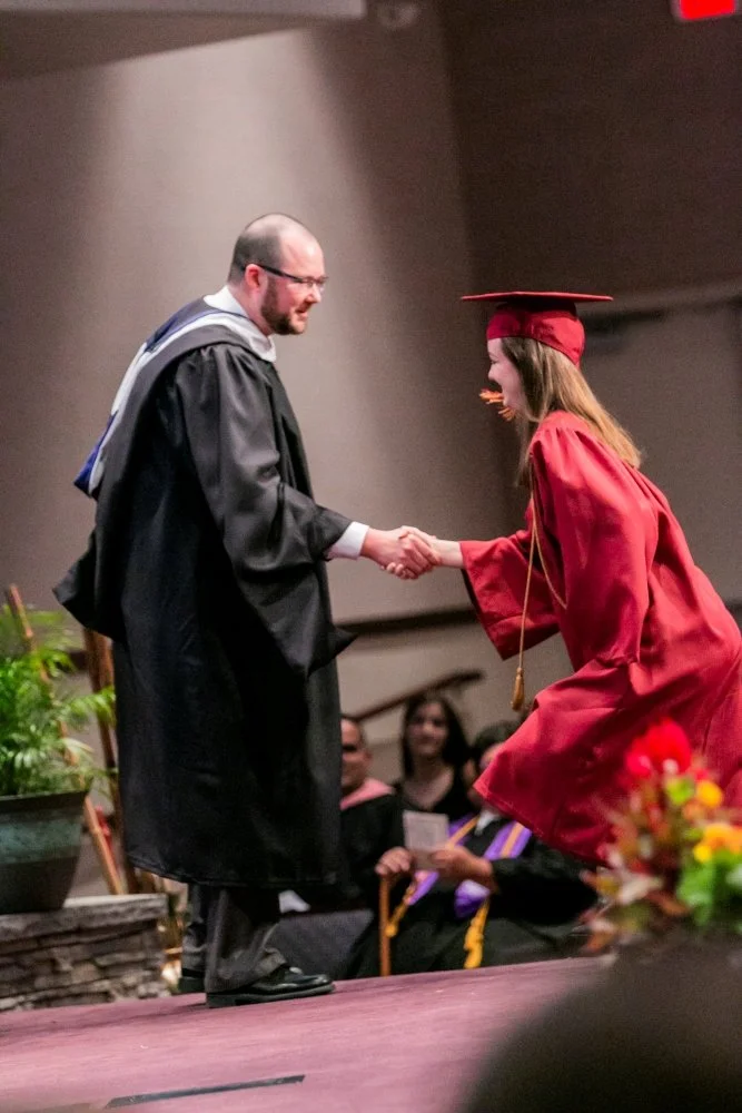 A graduate in a red cap and gown shaking hands with a man in an academic gown on stage during a graduation ceremony.