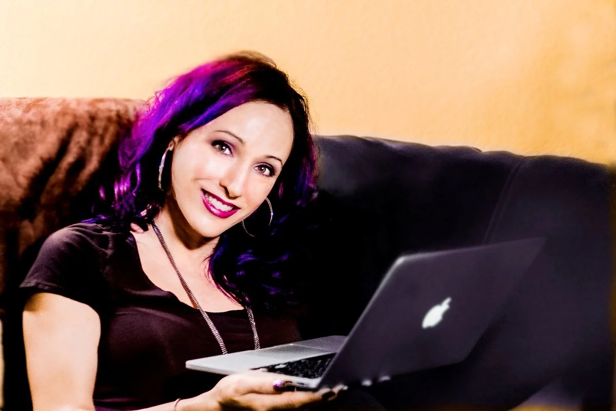 A woman with purple and black hair sitting on a couch, smiling at the camera, holding a MacBook laptop.